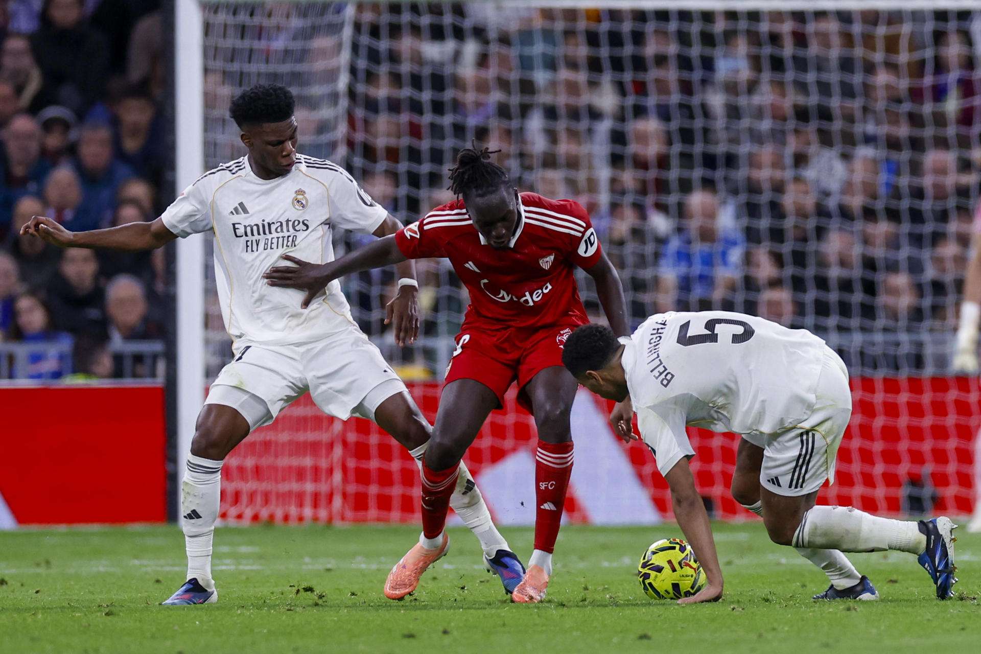 El centrocampista francés del Sevilla, Lucien Agoumé (c), disputa el balón ante los jugadores del Real Madrid, el francés Aurelien Tchouameni (i) y el inglés Jude Bellingham, durante el encuentro de la jornada 17 de LaLiga entre Real Madrid y Sevilla FC celebrado este sábado en el estadio Santiago Bernabéu, en Madrid. EFE / Javier Lizón.